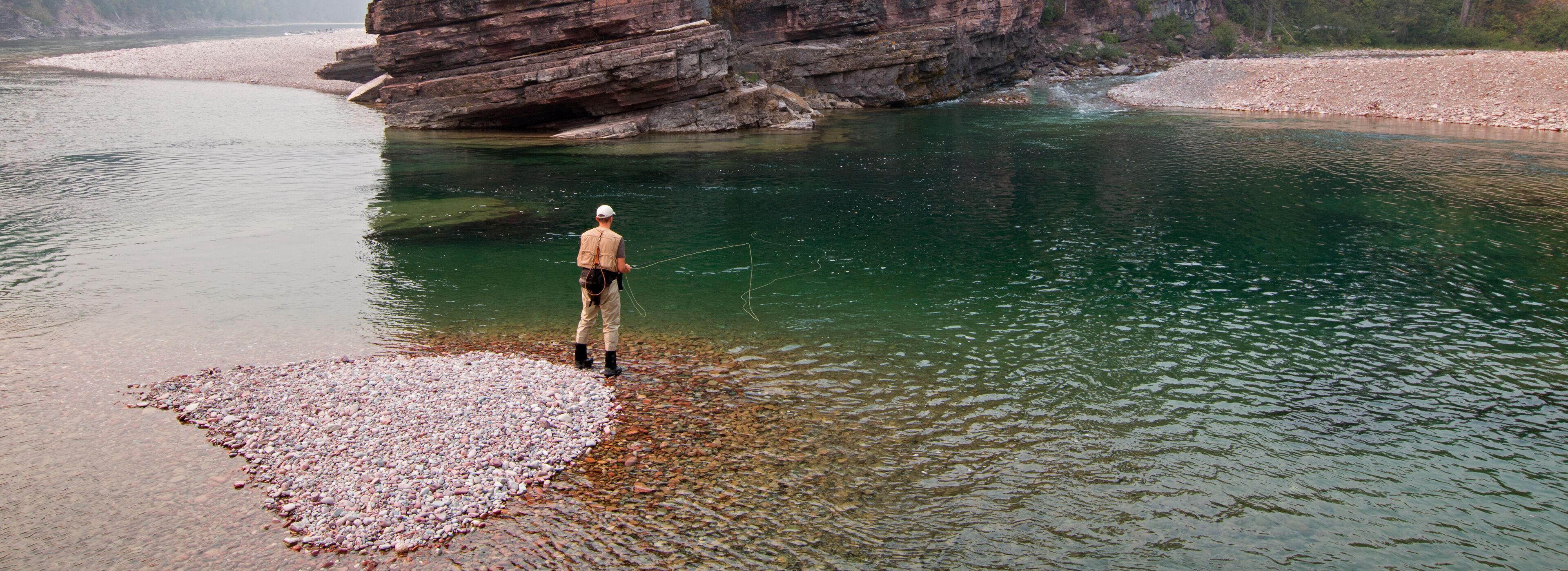 A person fishing by a clear river with rocky banks and a large rock formation in the background.