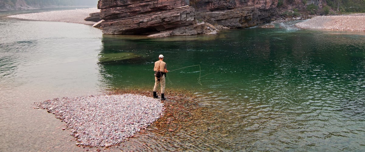A person fishing by a clear river with rocky banks and a large rock formation in the background.