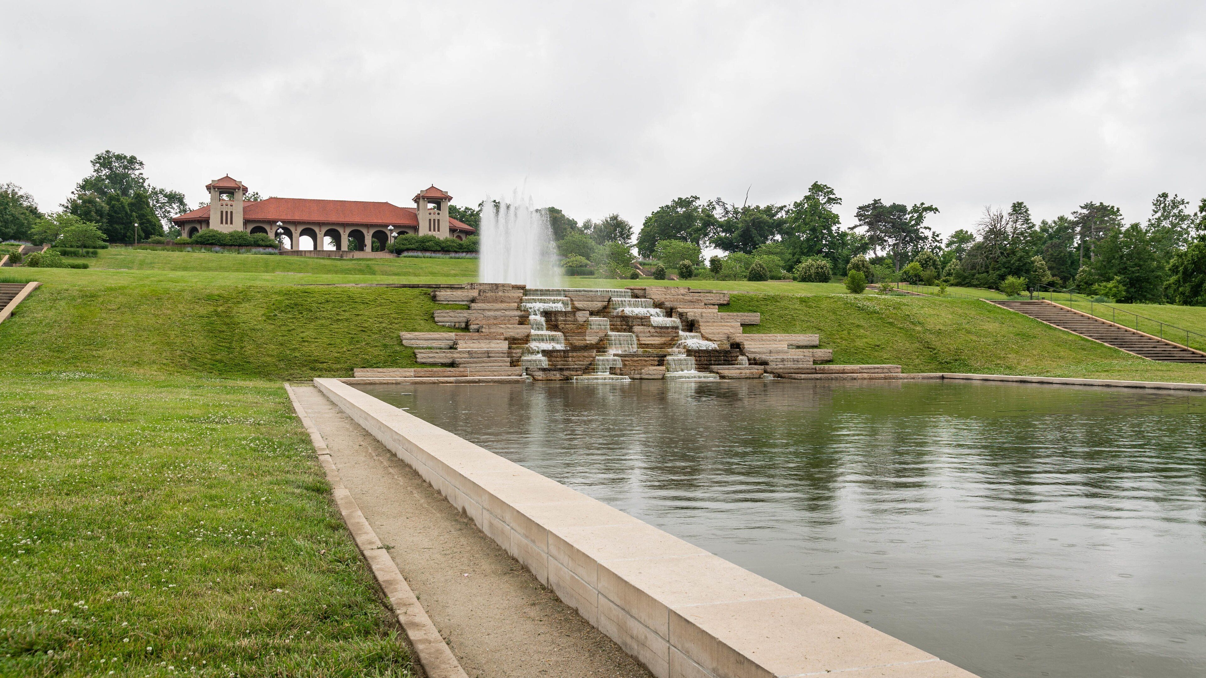 A stone fountain with water cascading down a tiered structure, surrounded by a pond and a grassy area with a building in the background.