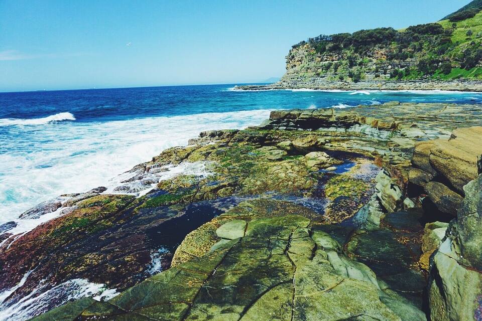 A rocky coastline with waves crashing against the shore and a cliff with vegetation in the background.