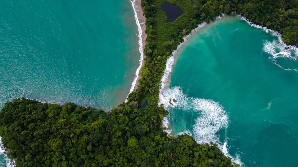 Vista aérea de una zona costera con aguas turquesas, una playa de arena, un bosque verde denso y un lago pequeño y oscuro.