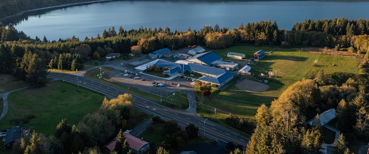 An aerial view of a lakeside industrial area with buildings, roads, and surrounding forested hills.