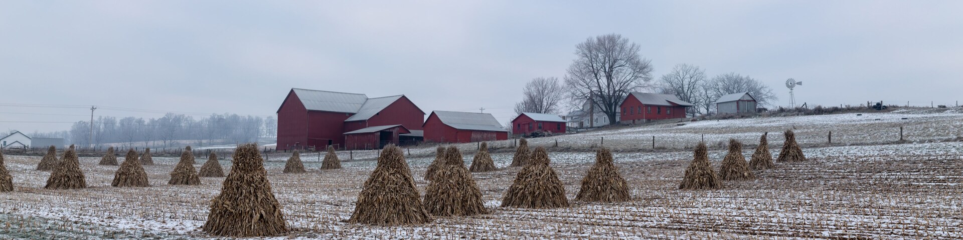 Corn Shocks in a Snowy Field with an Amish Farm with Red Buildings in the Background | Holmes County, Ohio