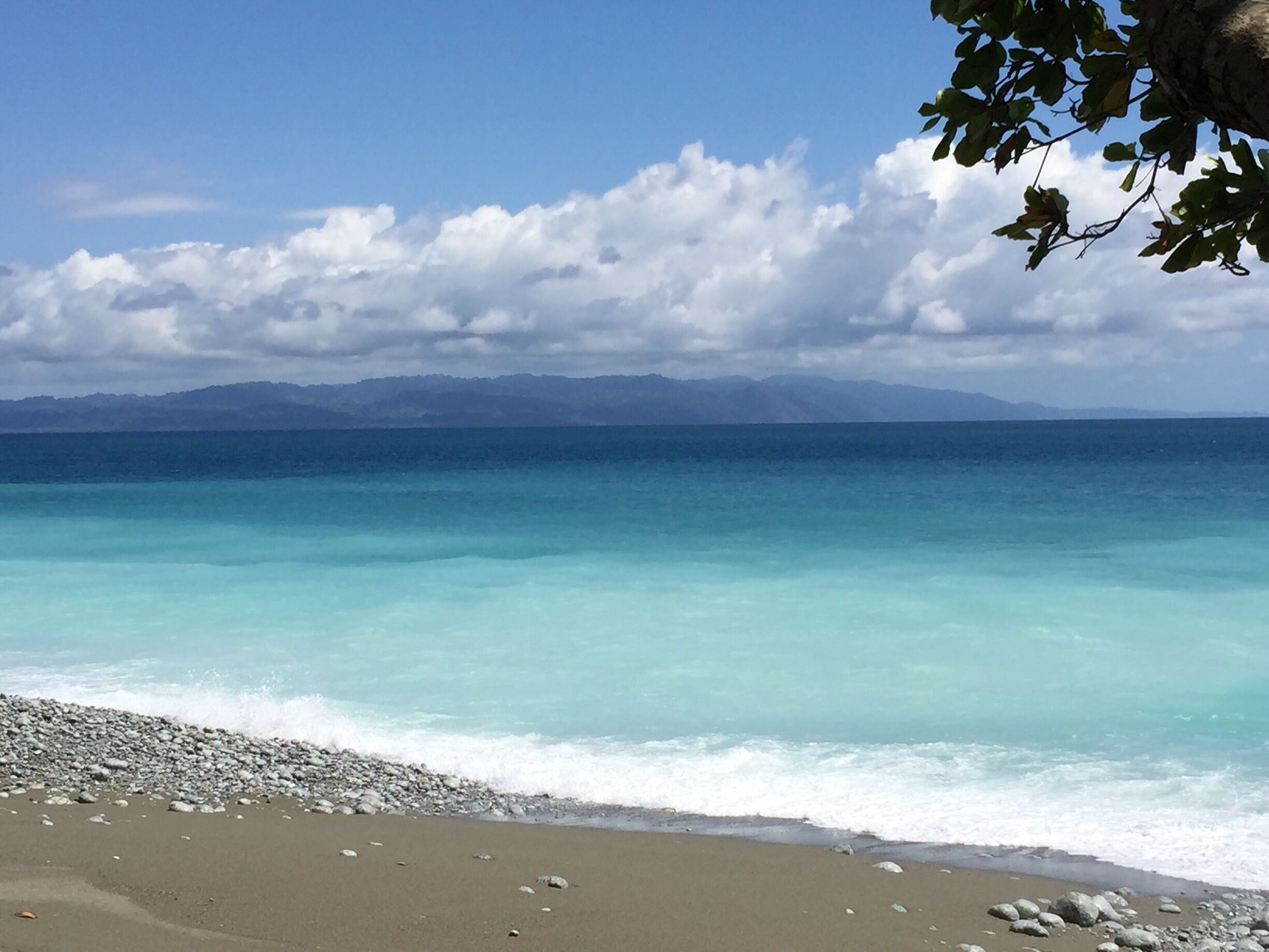 A beach with clear turquoise water, pebbles, and distant mountains under a partly cloudy sky.