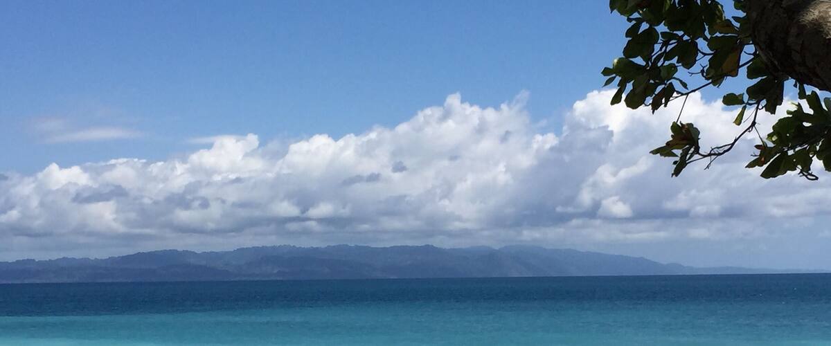 A beach with clear turquoise water, pebbles, and distant mountains under a partly cloudy sky.