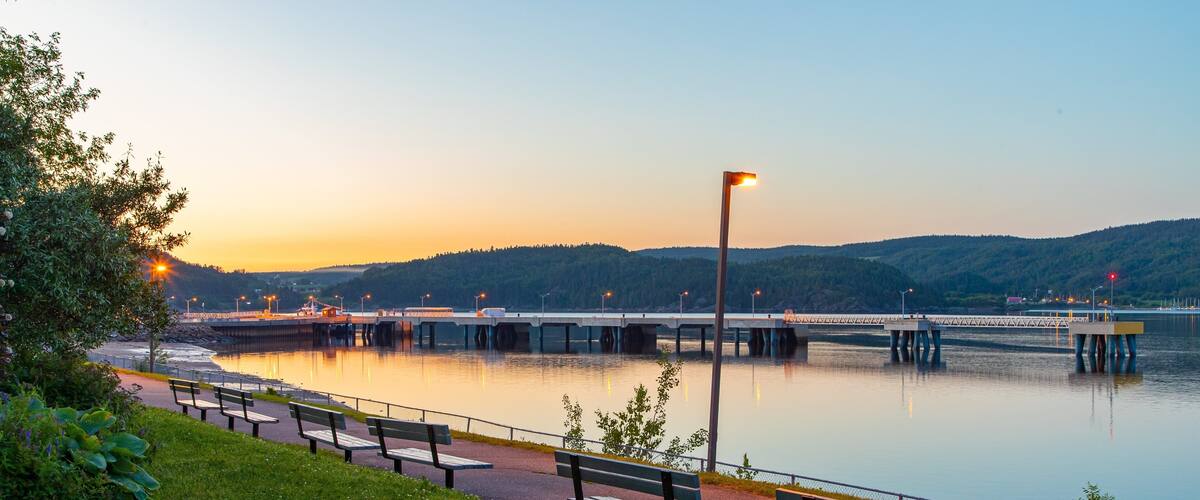 A riverside walkway with benches, a bridge, and a calm sunset.