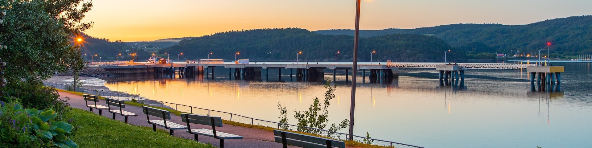 A riverside walkway with benches, a bridge, and a calm sunset.