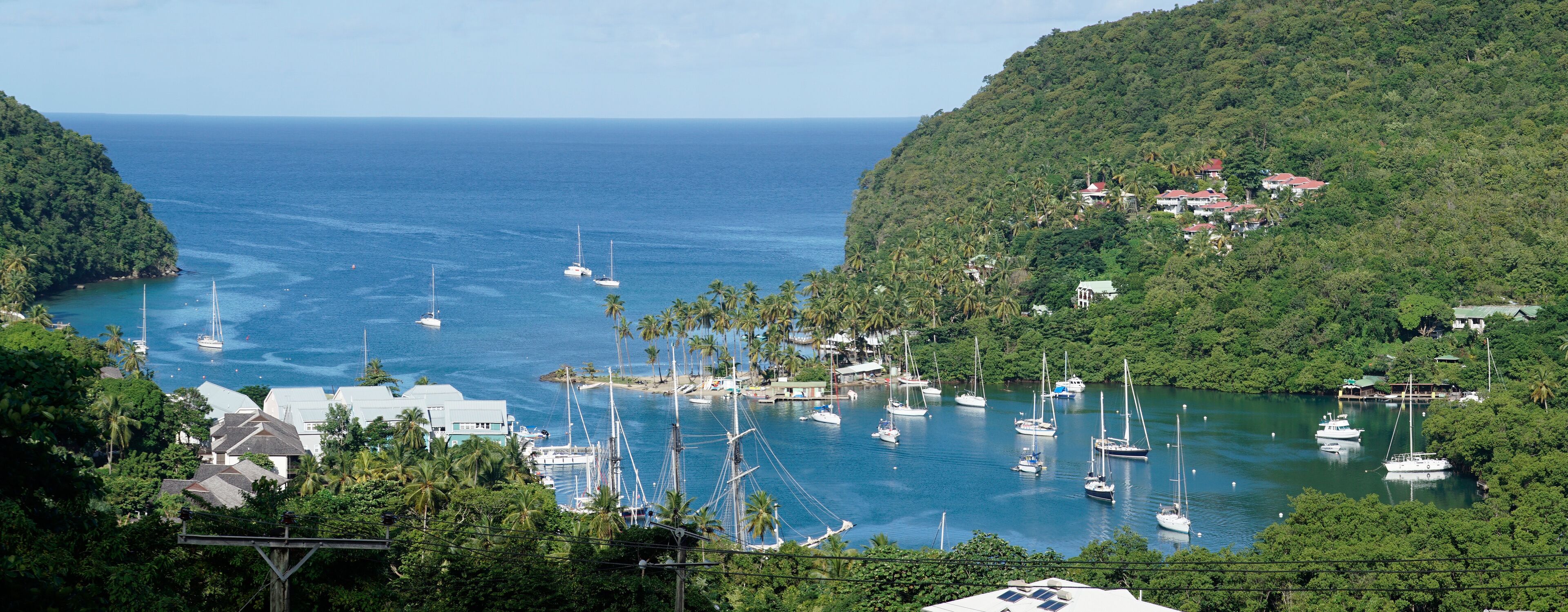 A coastal town with a marina filled with sailboats, surrounded by lush green hills and a clear blue sea.