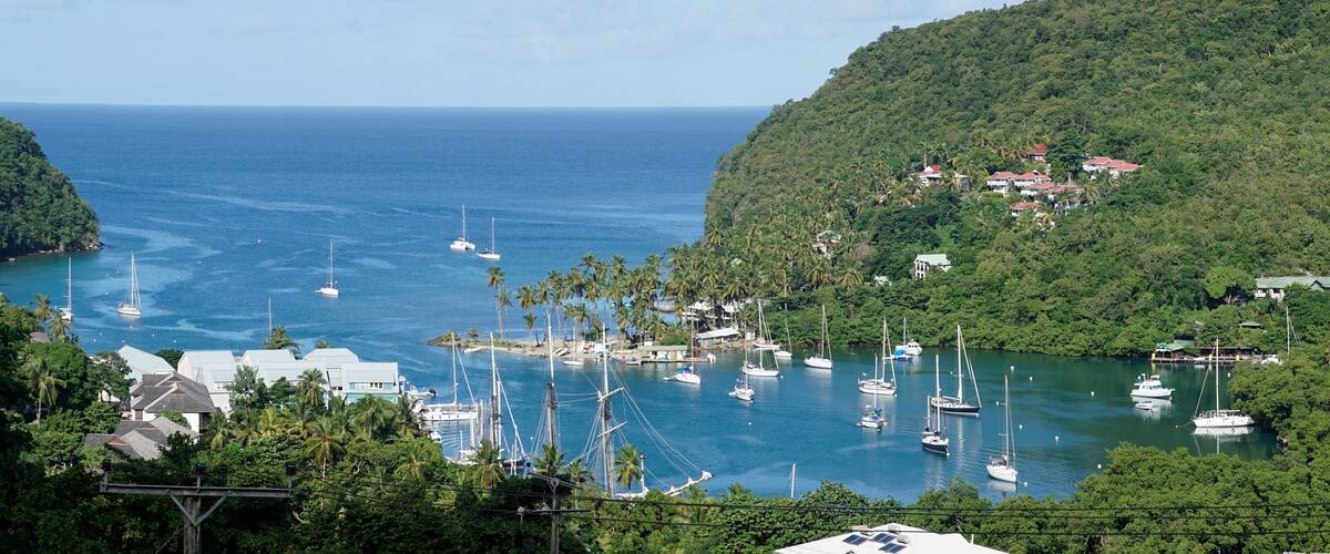A coastal town with a marina filled with sailboats, surrounded by lush green hills and a clear blue sea.