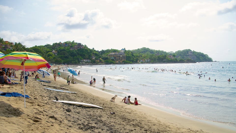 A crowded beach with people swimming and sunbathing, a colorful umbrella, and a building in the background.