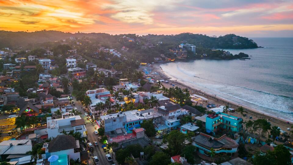 A coastal town with a beach, houses, and a hilly backdrop during sunset.