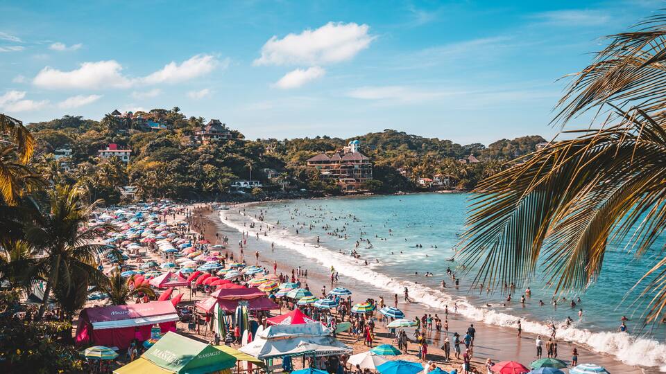 A crowded beach with colorful umbrellas and palm trees.