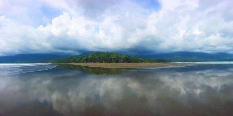 A beach with calm waters reflecting the sky and clouds, a sandy shore, and a distant forested hill.