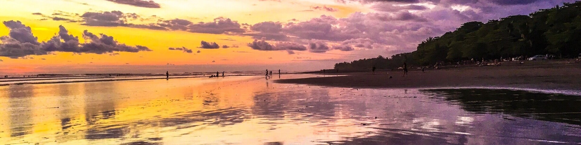 Un atardecer sobre una playa, con reflejos en la arena mojada y una colina boscosa al fondo.