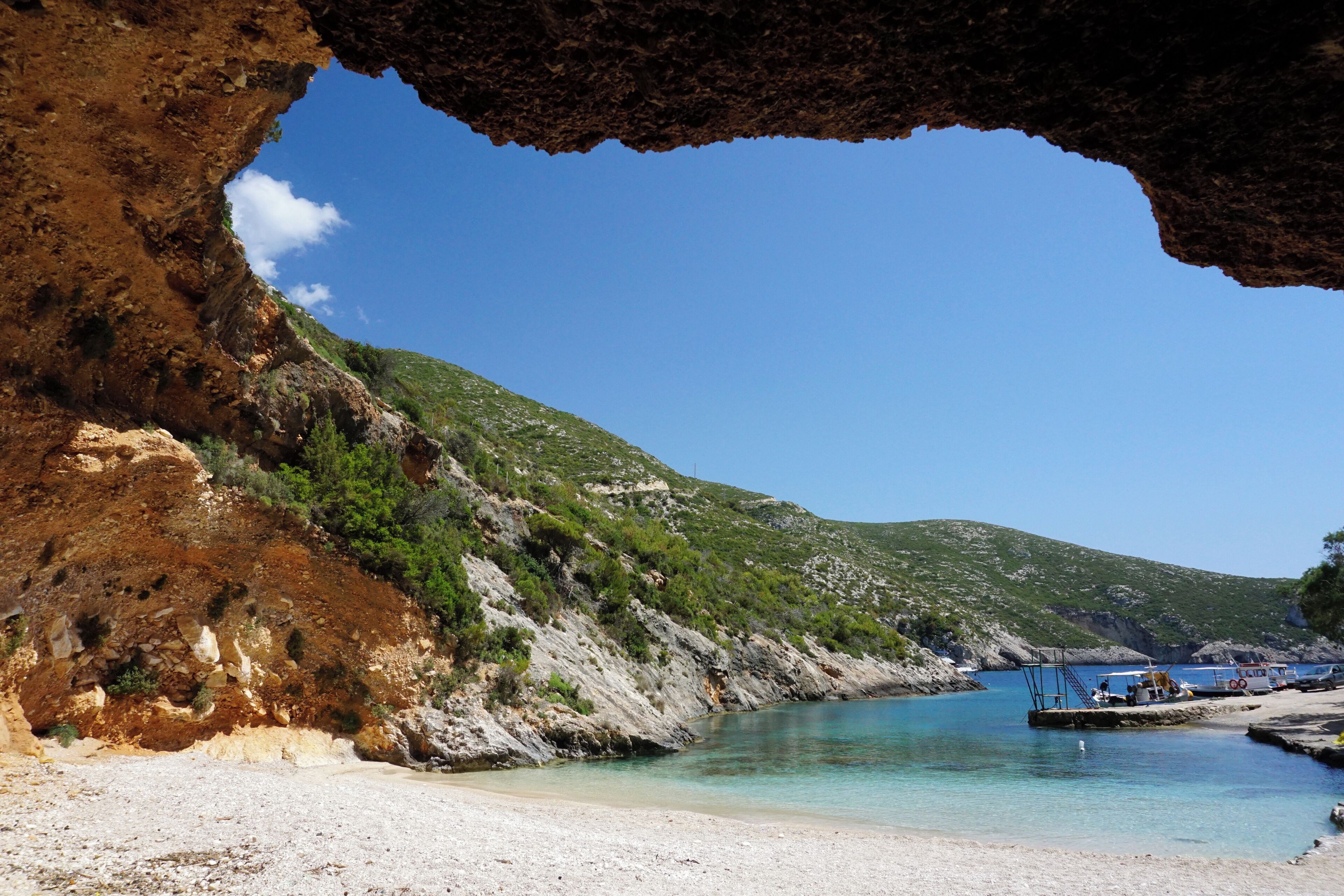 A coastal landscape with a clear blue sky, a rocky cliff, a sandy beach, and a calm sea with boats.