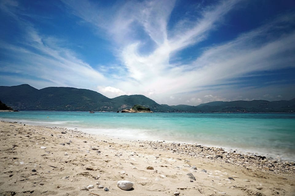 A beach with clear turquoise water, pebbly shore, and mountains in the background.
