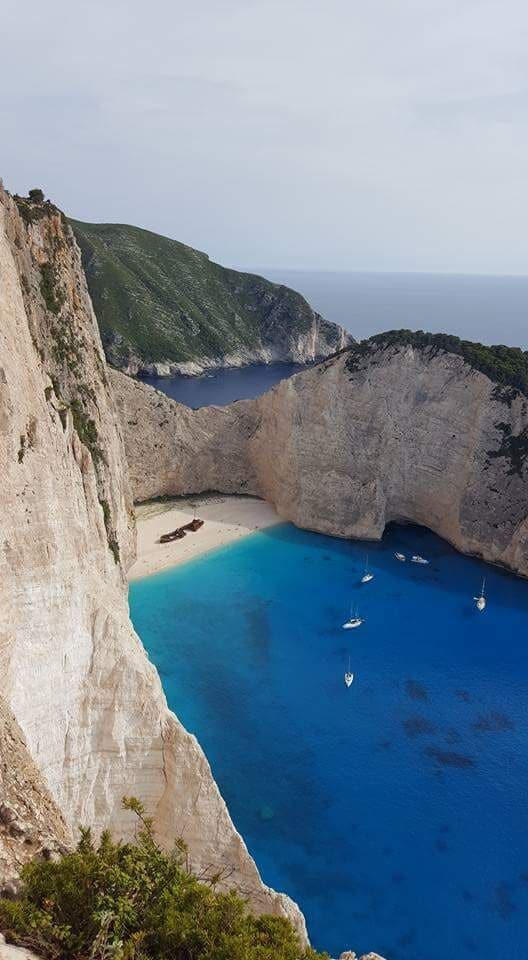 A coastal landscape with a prominent cliff, a sheltered bay, and several boats anchored in the clear blue waters.