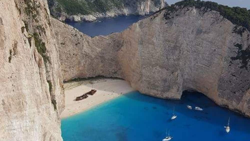 A coastal landscape with a prominent cliff, a sheltered bay, and several boats anchored in the clear blue waters.