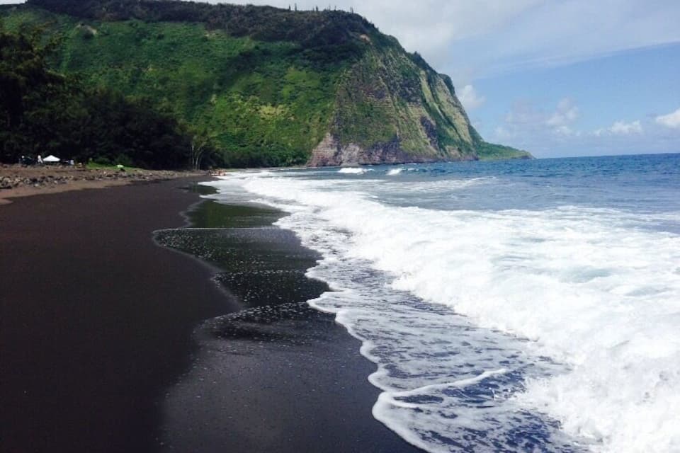 Una playa con arena oscura, olas rompiendo en la orilla y una ladera verde al fondo.