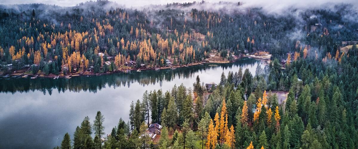 A lake surrounded by dense forests with autumn foliage, reflecting the trees and buildings on the water's surface.