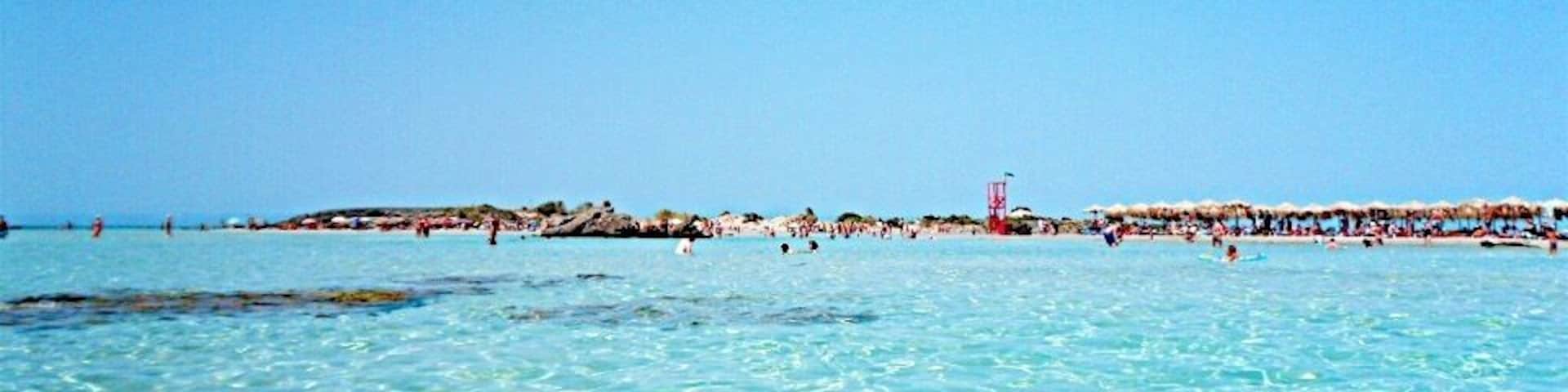 Água turquesa translúcida, uma praia de areia e um trecho de costa rochosa, com pessoas ao longe.