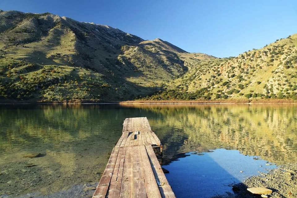 A wooden dock extending into a calm lake with clear reflections, surrounded by green hills and mountains.