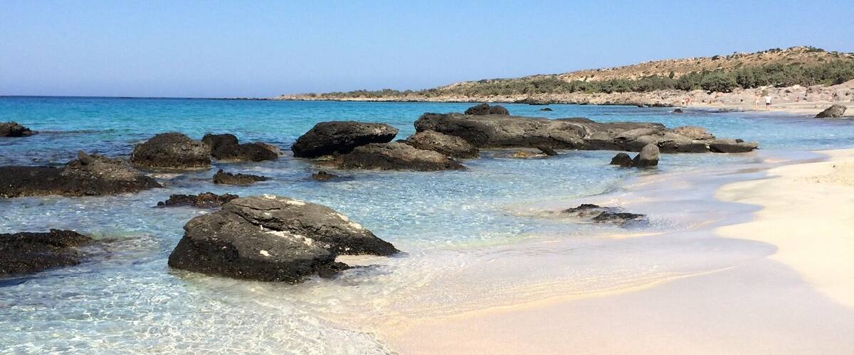 A beach with clear shallow waters, large rocks, and a sandy shore.