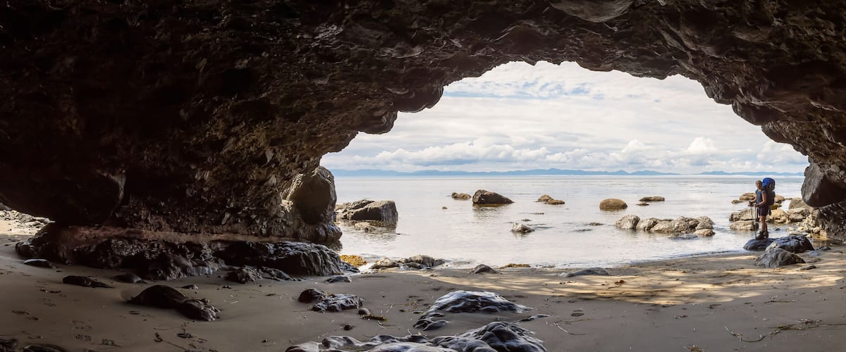 Une grotte rocheuse s’ouvrant sur une plage parsemée de rochers, avec une chaîne de montagnes visible au loin.