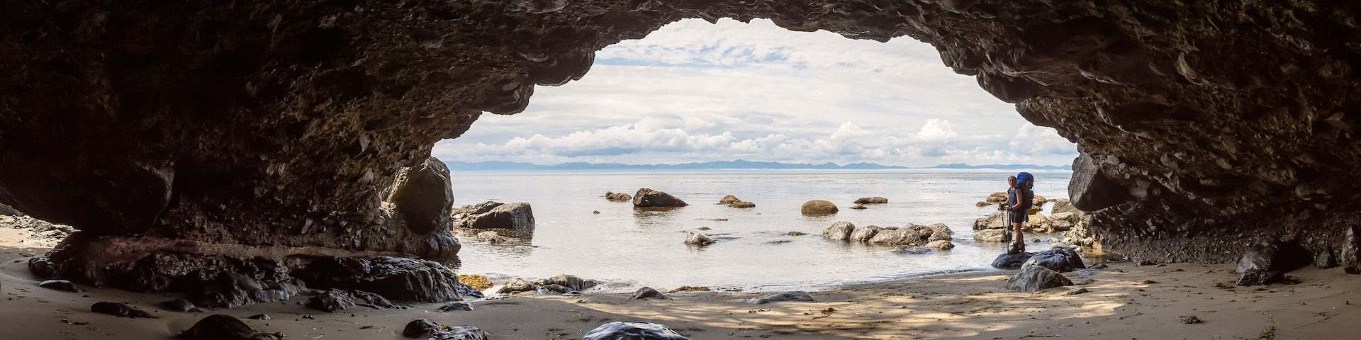 Boca de cueva rocosa que da a una playa con rocas dispersas y una cadena montañosa a lo lejos.