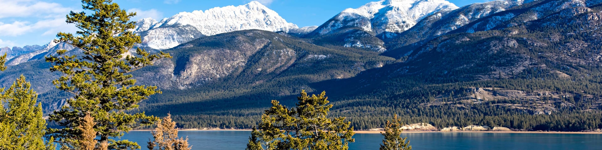 Paisagem montanhosa com picos nevados, céu azul límpido e um lago tranquilo rodeado por árvores de coníferas.