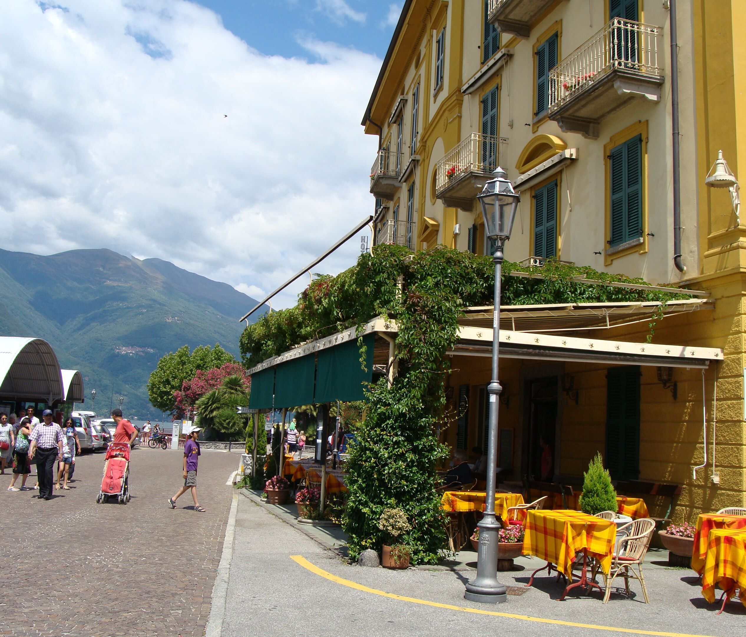 A cobblestone street with outdoor seating, a mountain in the background, and a yellow building with green shutters.