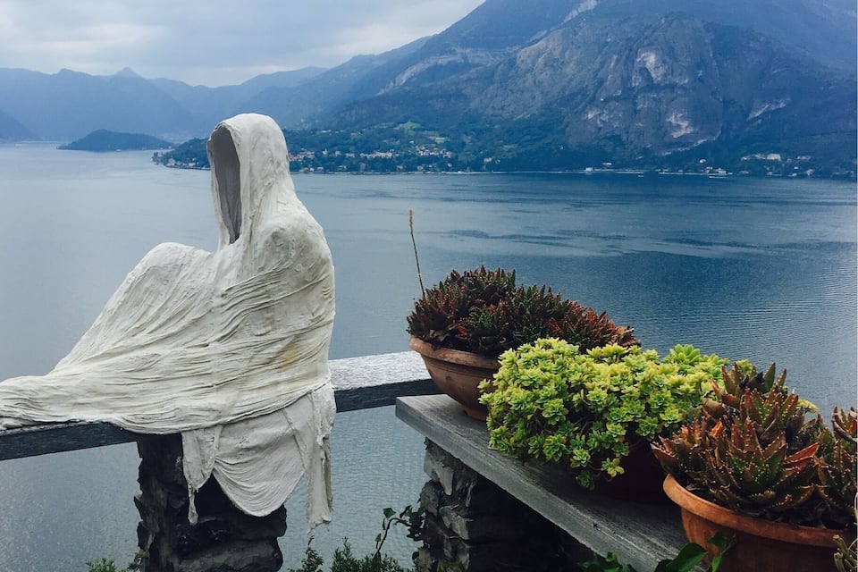 Una estatua de una figura cubierta con tela blanca, plantas en macetas y vistas a un lago rodeado de montañas.