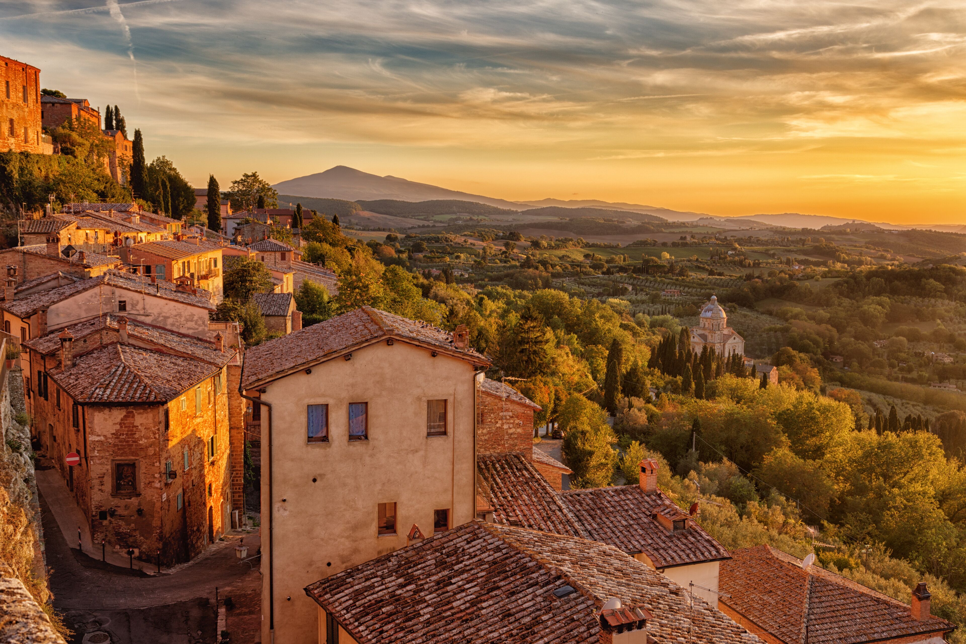 A historic town with terracotta-roofed buildings, a prominent church, and rolling hills in the background.