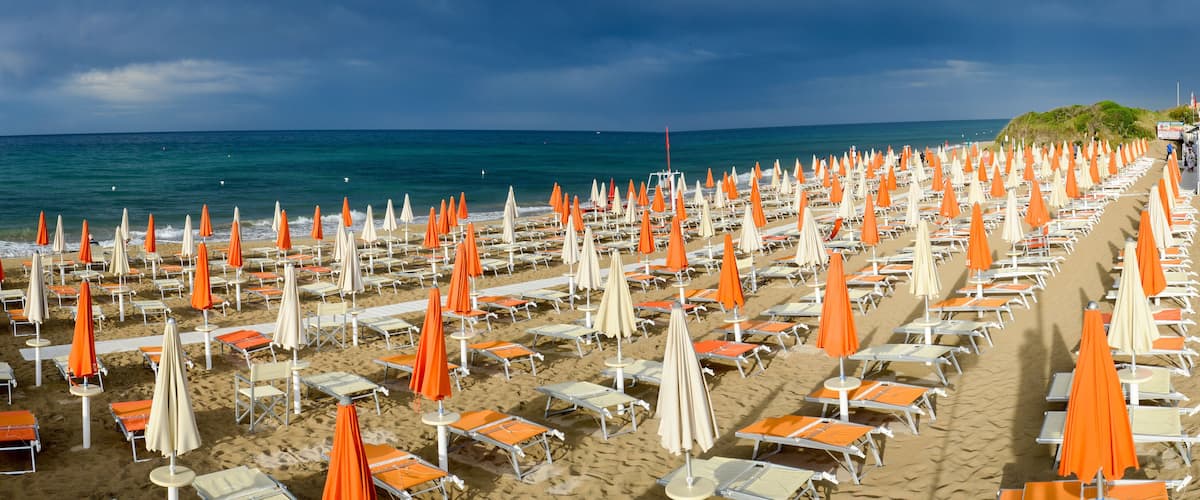 A beach with rows of sunbeds and umbrellas, a sandy shore, and the ocean in the background.