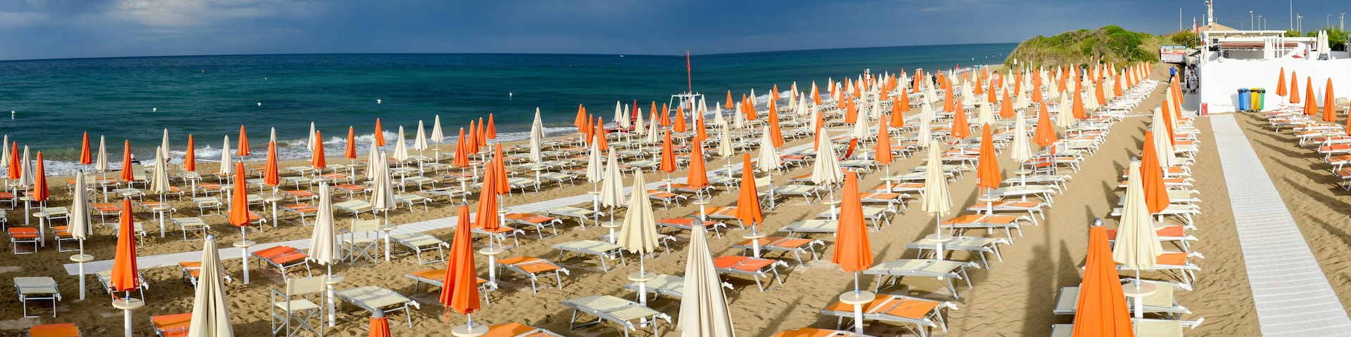 A beach with rows of sunbeds and umbrellas, a sandy shore, and the ocean in the background.