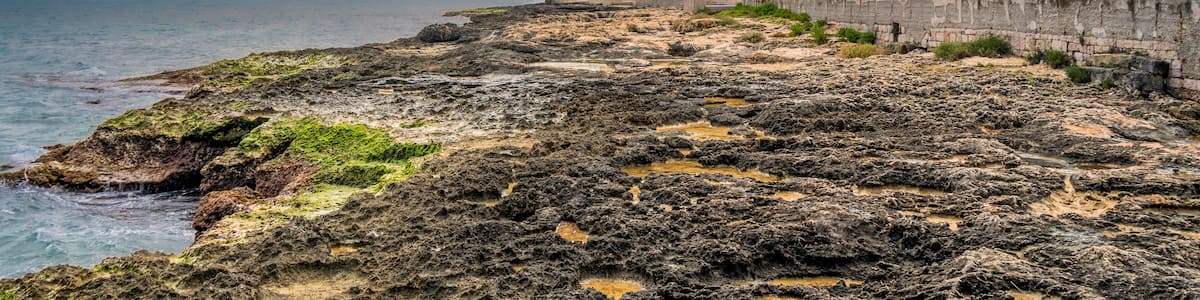 A rocky coastline with a lighthouse, a building with a yellow facade, and a clear sky with clouds.