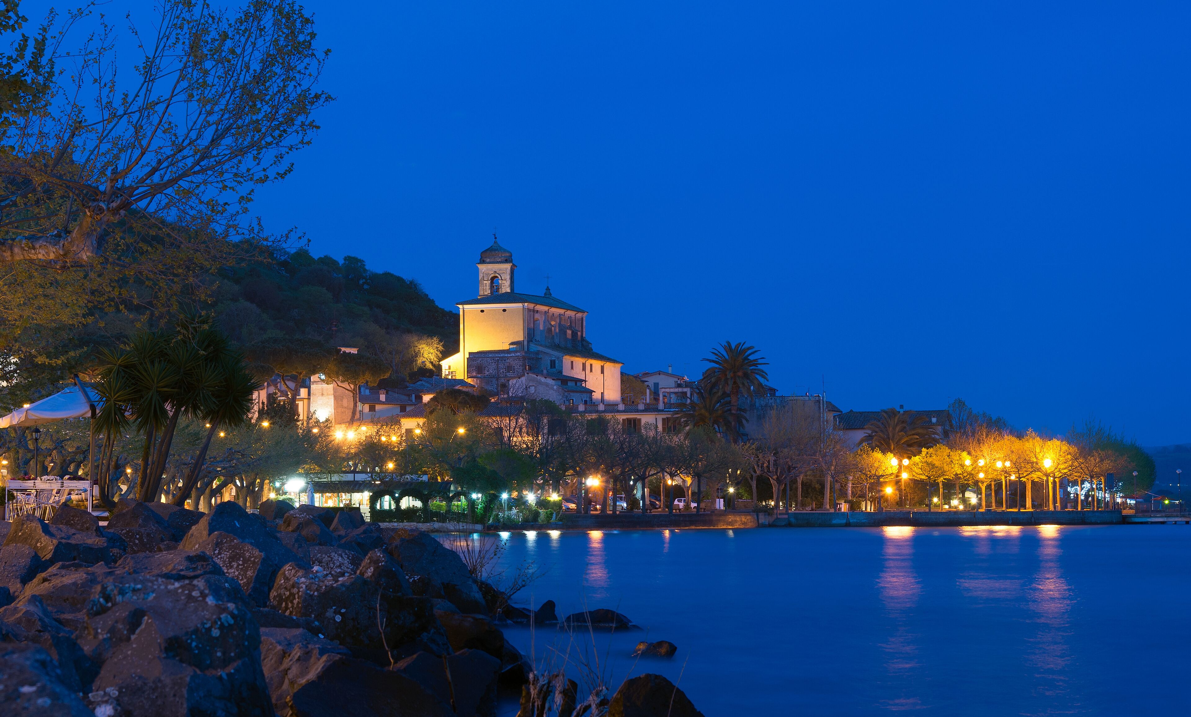 A coastal town at dusk with a prominent church on a hill, a calm sea, and a promenade with lights.