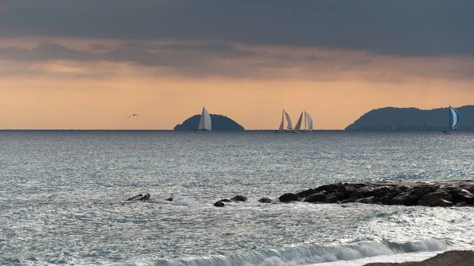 A coastal scene with a rocky shoreline, calm sea, and sailboats.