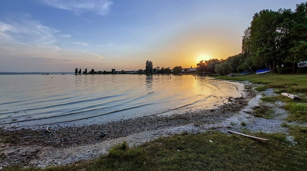 Un coucher de soleil sur un lac paisible, avec une rive de galets et des arbres sur la droite.