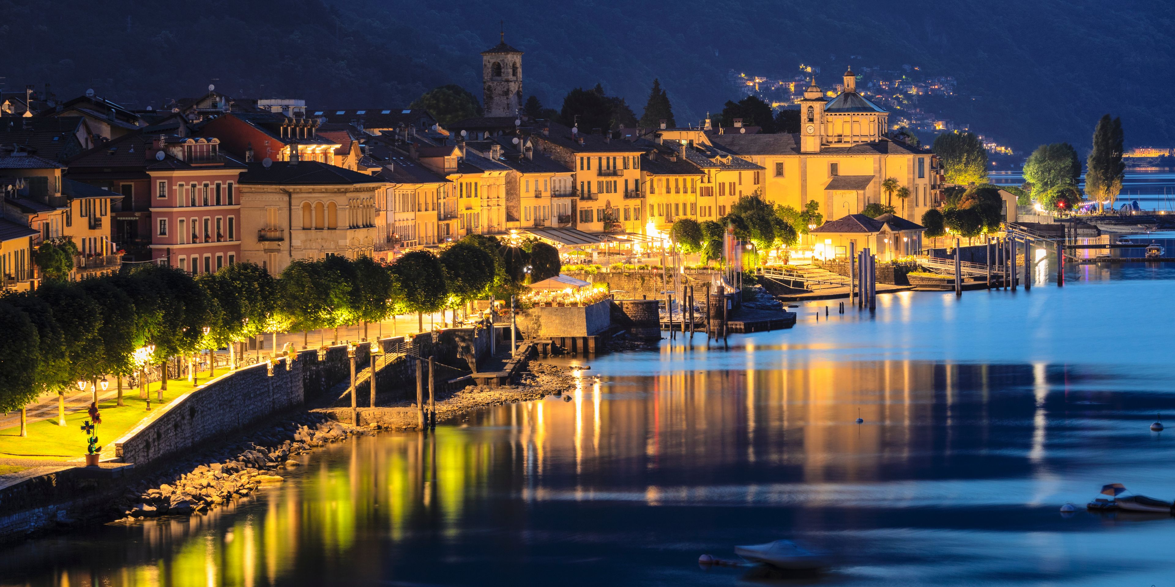 A riverside town at night with illuminated buildings, a bridge, and a calm water surface reflecting the lights.
