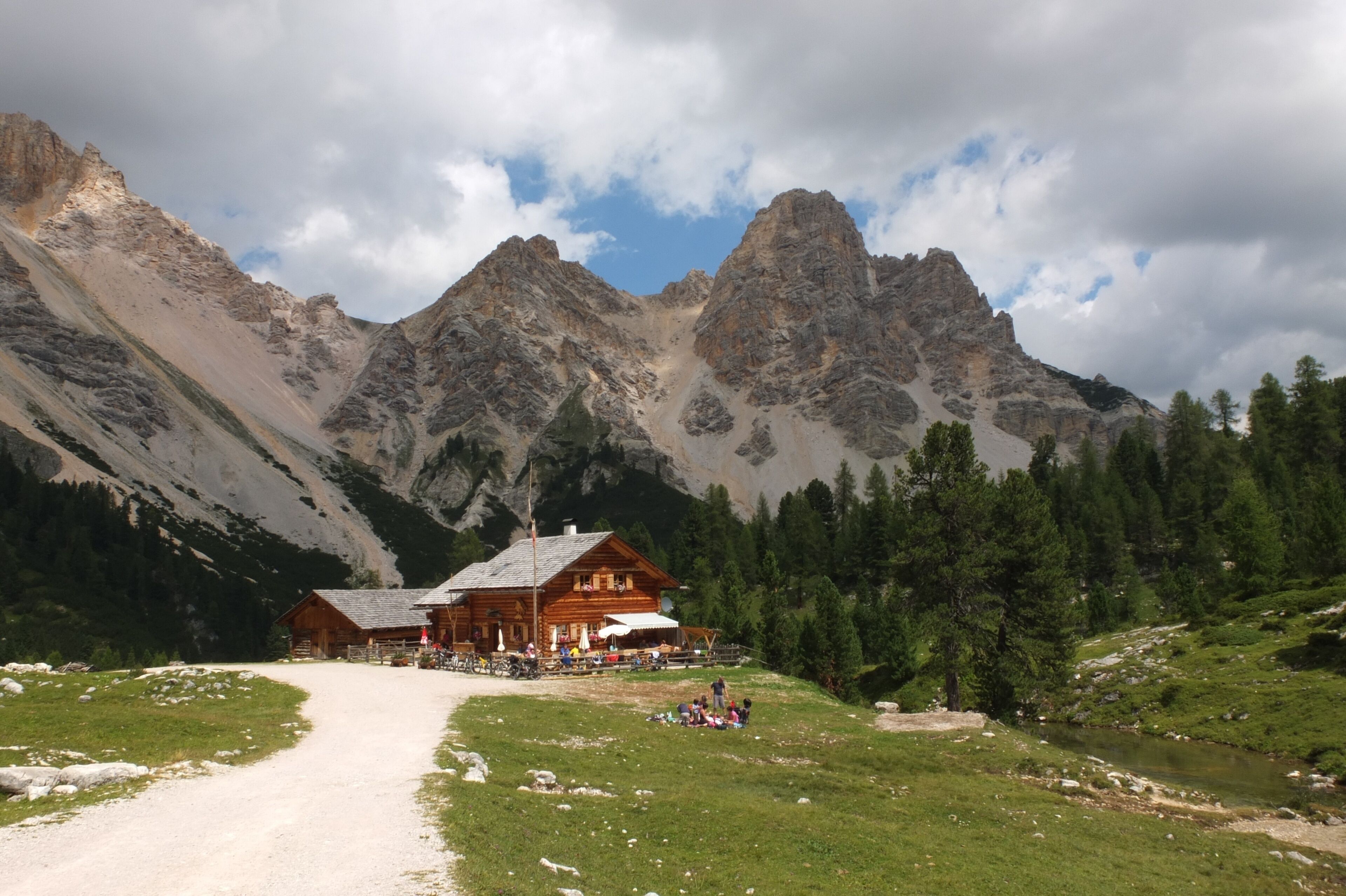 A mountain lodge with a dirt path leading to it, surrounded by forest and rocky peaks.