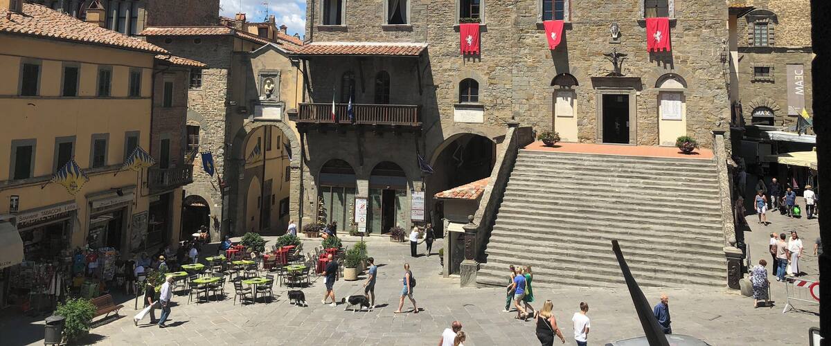 A historic square with a prominent clock tower, stone buildings, and outdoor seating areas.