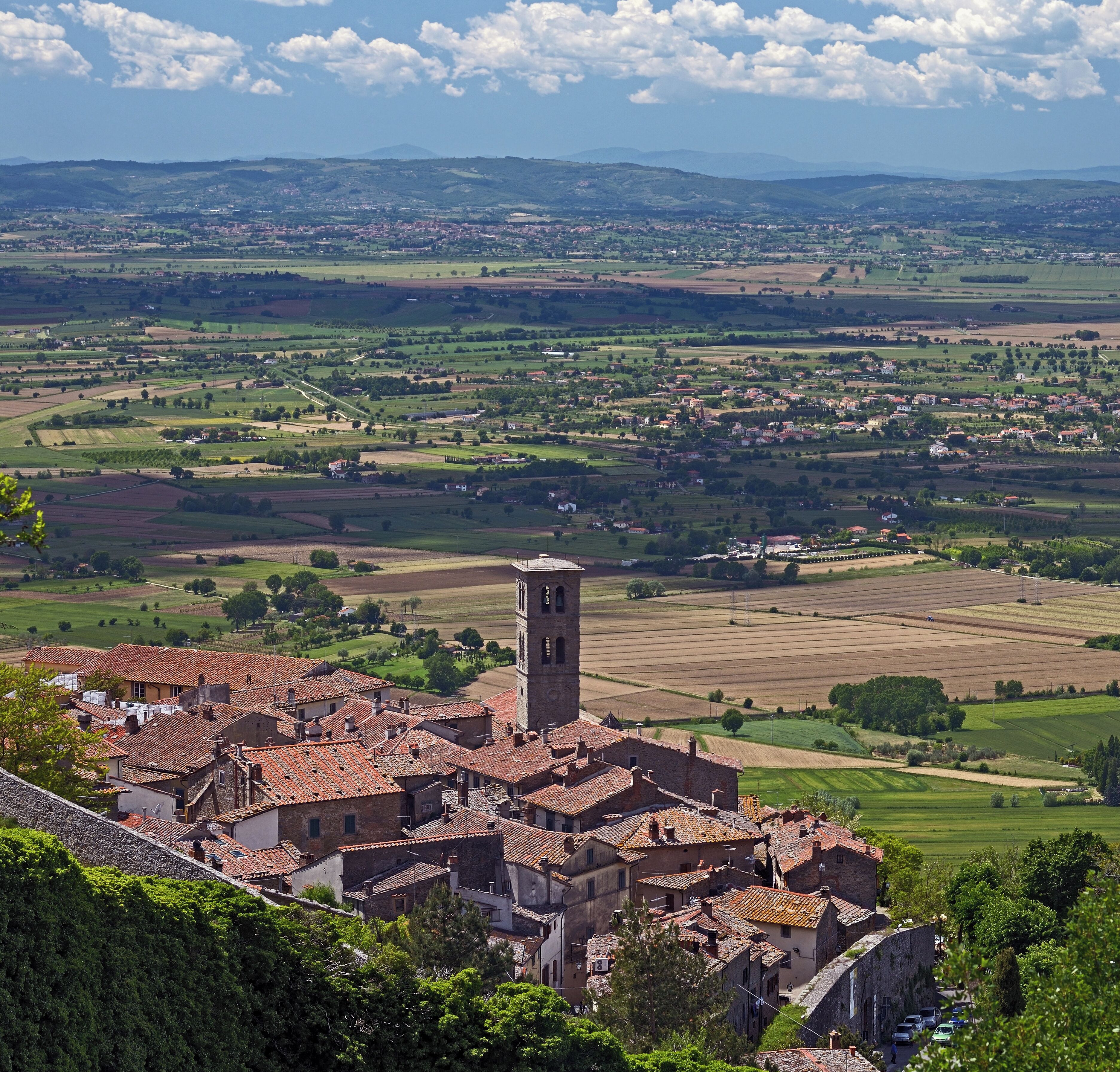 A village with terracotta-roofed buildings, a prominent tower, and expansive green fields.