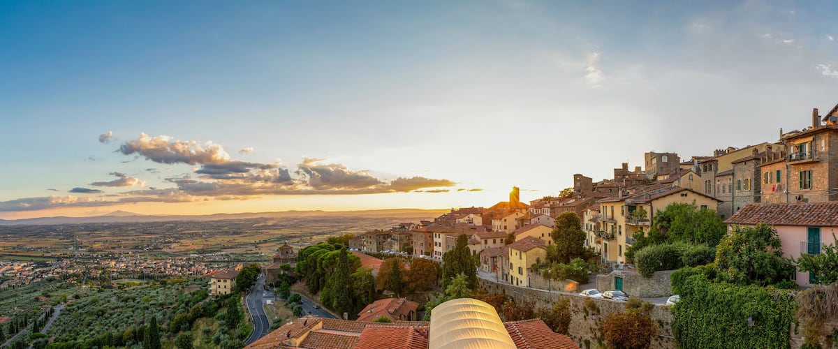 Une ville perchée sur une colline, avec des bâtiments aux toits de tuiles rouges, une route sinueuse et une vue lointaine sur une vallée.