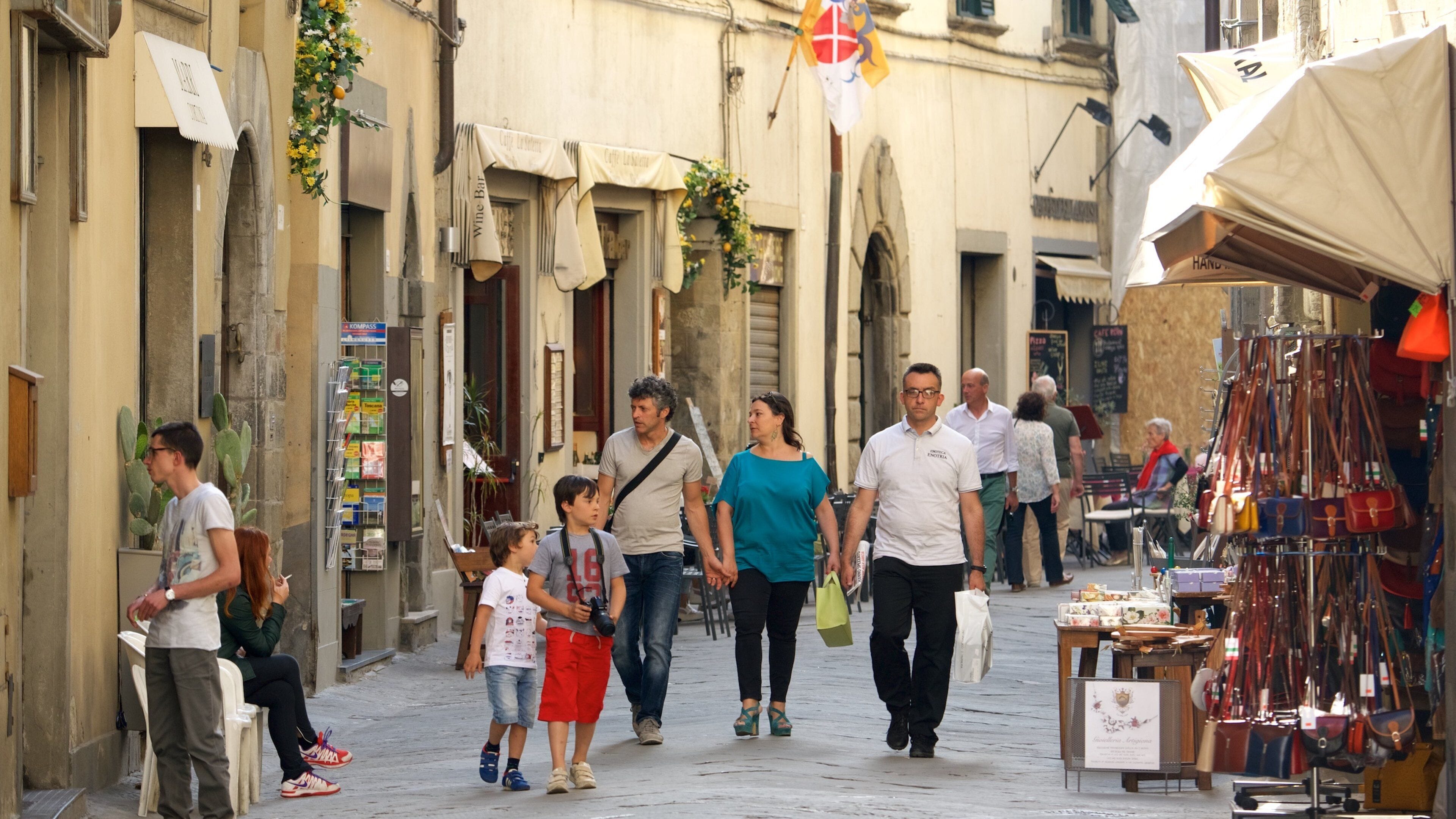 Rua movimentada com pessoas caminhando, uma banca de mercado com sacolas e um edifício com uma placa.