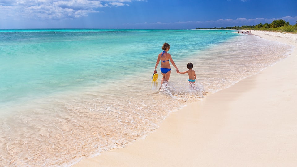 A woman and a child walking on a sandy beach with clear turquoise water.