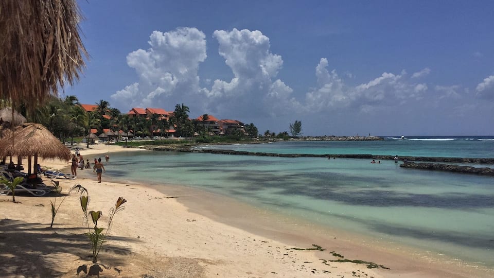 A tropical beach with clear turquoise waters, palm trees, and a resort in the background.