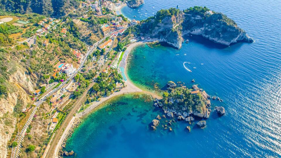 An aerial view of a coastal town with a clear blue sea, rocky outcrops, and a winding road.