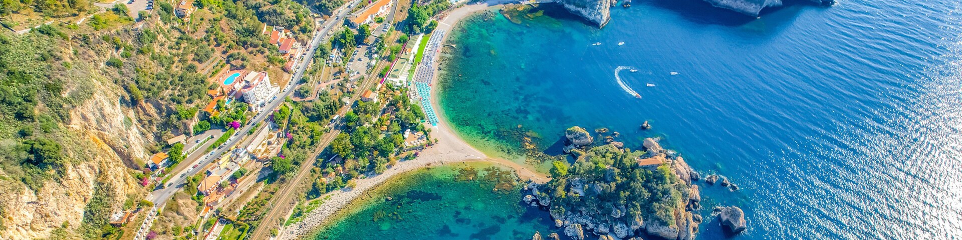 Une vue aĂ©rienne dâune ville cĂŽtiĂšre, avec une mer bleue et limpide, des affleurements rocheux et une route sinueuse.