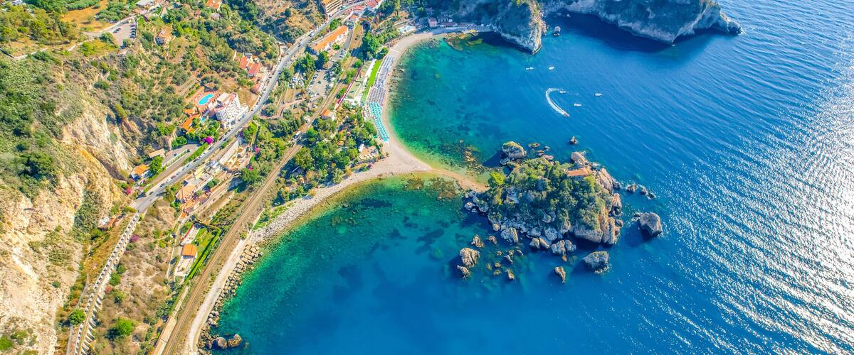 An aerial view of a coastal town with a clear blue sea, rocky outcrops, and a winding road.
